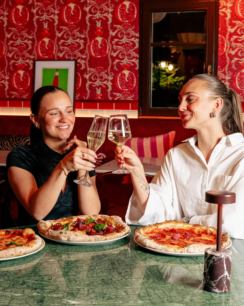 Deux amies trinquent autour de pizzas napolitaines dans la salle rouge de Scopertino